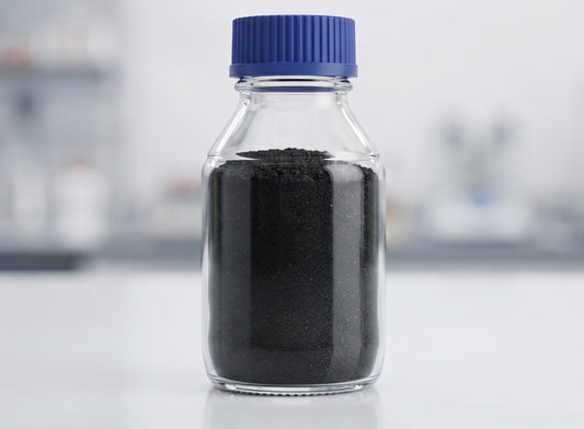A clear glass bottle with a blue ribbed cap filled with black powder, set against a blurred light gray laboratory background.